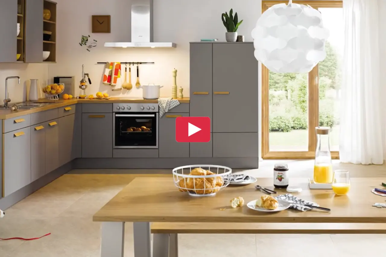 Moderne Küche in Grau mit Holzarbeitsplatte, Backofen und großer Fensterfront. Im Vordergrund ein Esstisch mit Frühstück und Getränken. Helles Küchendesign mit warmen Akzenten für gemütliches Kochen und Wohnen.